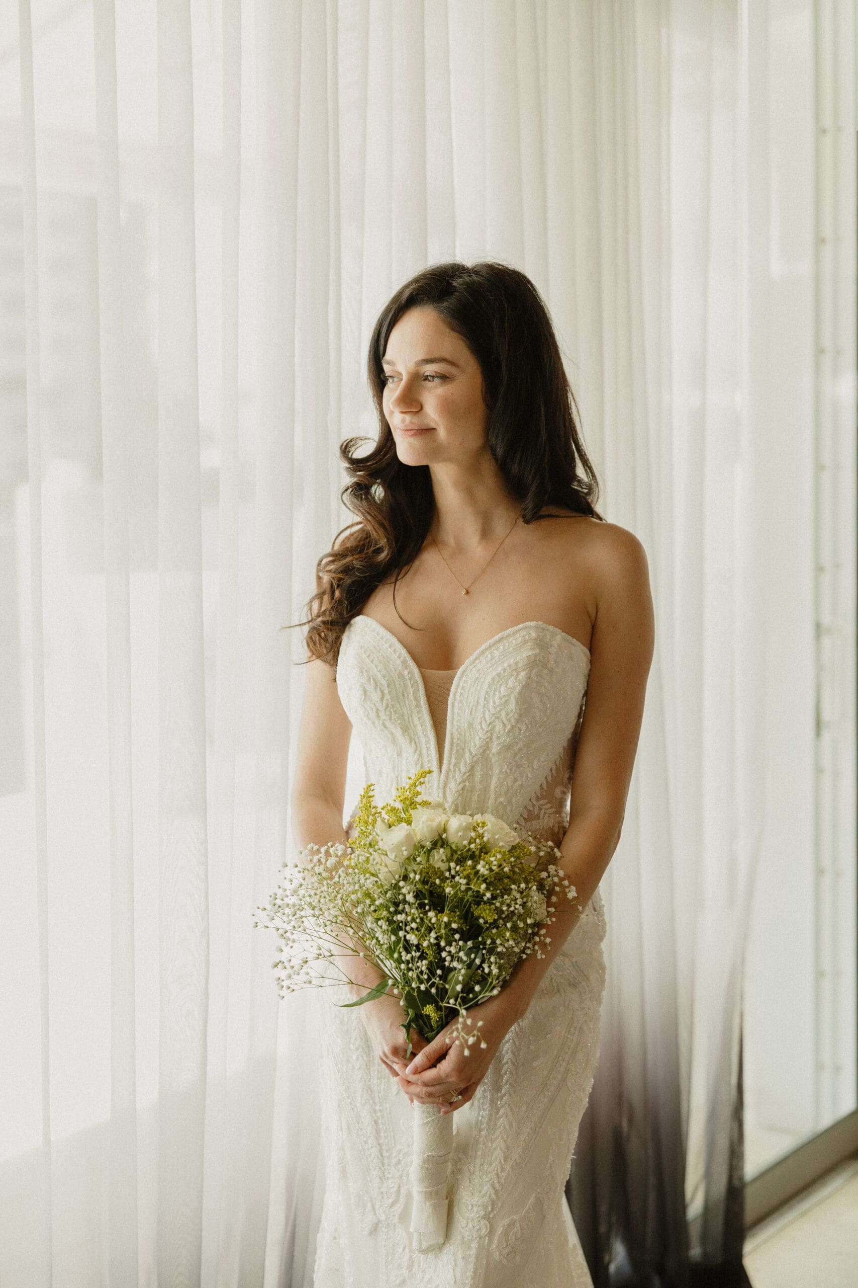 Bride in strapless lace gown holding white rose bouquet looking out window at Grateful Palate Fort Lauderdale wedding reception